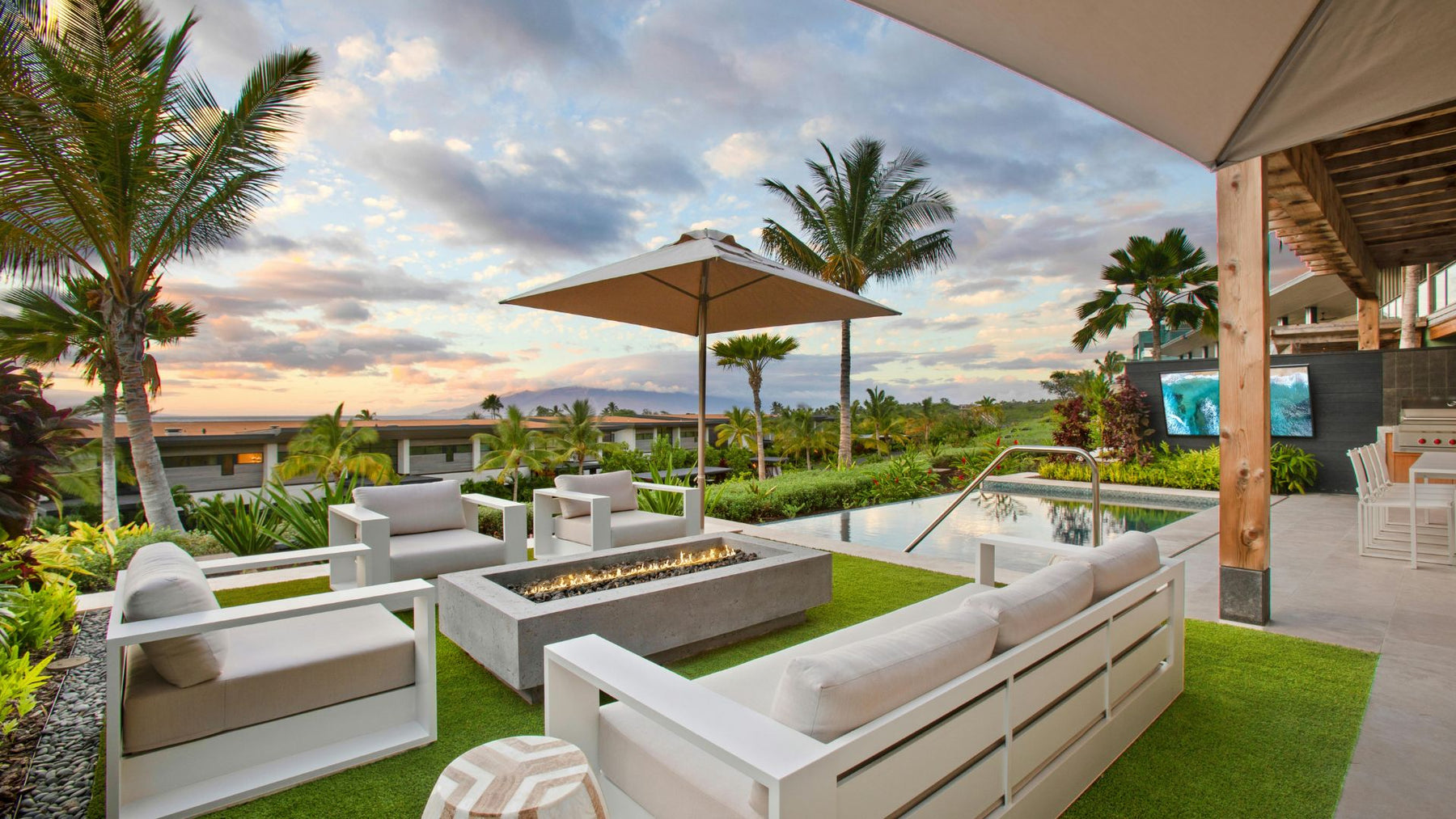 Luxury Airbnb outdoor space with modern fire pit, lounge seating, infinity pool, and tropical landscaping at sunset"